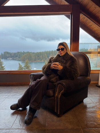 Una mujer con abrigo de piel, gafas de sol y vincha negra sostiene una taza sentada en un sillón frente a un ventanal