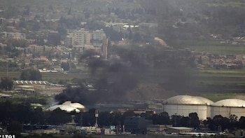 Vista aérea de una refinería de petróleo con humo negro elevándose sobre un tanque. Se observan edificios y zonas verdes en el fondo