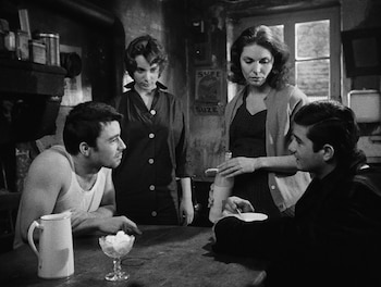 Imagen en blanco y negro de cuatro personas en una habitación. Dos hombres sentados a una mesa de madera, con un jarro y un vaso. Dos mujeres de pie detrás de ellos