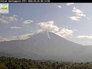 Imagen de cámara web del Volcán Santiaguito con una columna de humo o ceniza ascendiendo desde su cráter. Bosques en primer plano, cielo con nubes