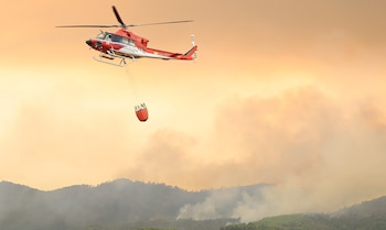 Helicóptero rojo y blanco con texto Gobierno de España y Avincis Iberia, balde de agua, sobre montañas con humo y cielo anaranjado