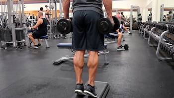 Hombre de espaldas en un gimnasio realiza ejercicio de pantorrillas sobre un step con mancuernas, sus talones están fuera del borde, otras personas y máquinas al fondo.