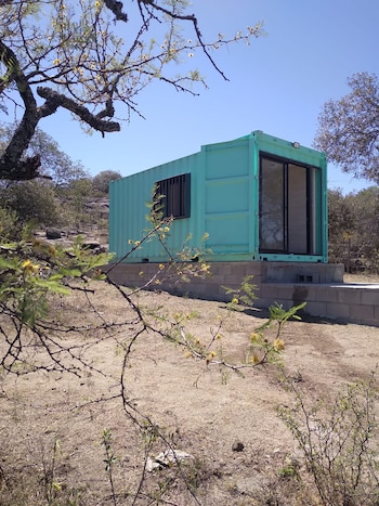 Una mini casa prefabricada de color turquesa sobre una base de hormigón en un paisaje seco con árboles y arbustos dispersos bajo un cielo azul claro