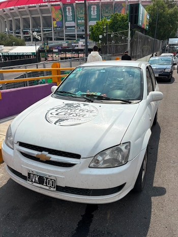 Un auto Chevrolet blanco con calcomanía "América en Corsa" y matrícula argentina está estacionado frente a un estadio con banderas del Mundial 2026