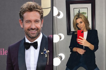 Hombre de traje con pajarita sonríe; mujer de blazer negro y camiseta blanca se toma una selfi con teléfono rojo frente a un espejo con luces y un cuadro.