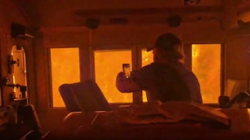 Una persona de espaldas con gorra sostiene un teléfono móvil, apuntando hacia ventanas que muestran intensa luz anaranjada. Se observan muebles en el interior oscuro