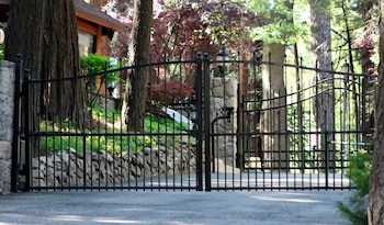 Un gran portón de hierro forjado negro, parcialmente abierto, en una entrada arbolada que lleva a una propiedad privada con pilares de piedra y abundante vegetación