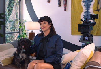 Una mujer de cabello oscuro con una chaqueta vaquera y un perro Poodle cruzado negro y blanco están sentados en un sofá marrón junto a una cámara