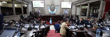 Amplia sala de un parlamento con muchos diputados sentados en escritorios, pantallas electrónicas y banderas nacionales
