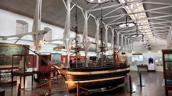 Interior de museo con una gran maqueta de barco de vela, vitrinas, cuadros, paneles informativos y techo con vigas de madera