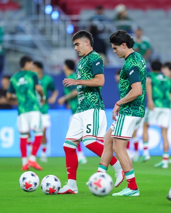 Dos futbolistas vestidos con camiseta verde y pantalón corto blanco manejan balones sobre el césped