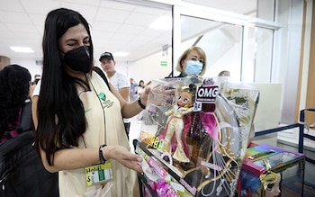 Una mujer con cabello oscuro, mascarilla negra y chaleco beige sostiene una muñeca L.O.L. Surprise! OMG en su empaque. Otros individuos aparecen borrosos en el fondo de un interior
