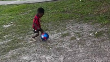 Un niño con camiseta roja y negra corre mientras patea una pelota sobre un terreno de césped y tierra