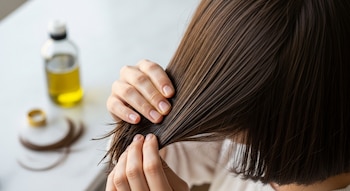 Primer plano de unas manos aplicando aceite en las puntas de un cabello castaño oscuro. En el fondo, una botella borrosa de aceite de oliva sobre una superficie blanca.