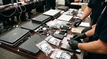 Varias computadoras portátiles, teléfonos móviles, cables en bolsas y discos externos sobre una mesa examinados por agentes con guantes negros.