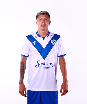 Hombre joven con camiseta de fútbol blanca y azul, escudo del club, logo de Saphirus, logo de Bajaj, y pantalón azul, con tatuaje en brazo izquierdo