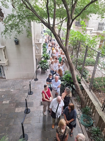 Vista superior de una larga fila de personas esperando en un sendero de piedra. Un gran árbol con follaje verde y edificios clásicos flanquean el área