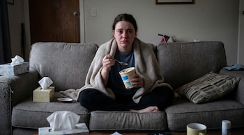 Mujer sentada en un sofá con una manta, sostiene un recipiente de helado y una cuchara, con lágrimas en los ojos, rodeada de cajas de pañuelos.
