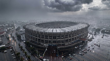 Vista aérea del Estadio Azteca con una fuerte lluvia. Se ven nubes oscuras, el estadio, calles mojadas con autos y varios estacionamientos.