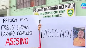 Dos pancartas de protesta con texto y la foto de un hombre, sostenidas frente a una señal de la Policía Nacional del Perú en una edificación