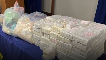 Una mesa cubierta con un mantel azul exhibe una gran pila de paquetes rectangulares blancos y dos bolsas transparentes llenas de artículos irregulares