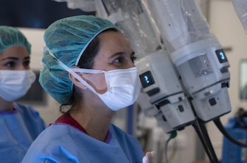Una cirujana con mascarilla y gorro quirúrgico observa junto a los brazos mecánicos de un robot de operaciones