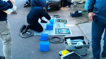 Agentes policiales examinan paquetes azules colocados sobre el asfalto junto a maletines periciales y carteles oficiales