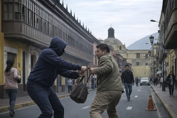 Dos hombres tiran de un bolso en una calle. Uno usa capucha, el otro chaqueta. Hay edificios con balcones, peatones, una cúpula y una montaña al fondo.