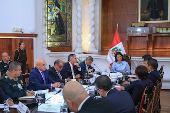 Varias personas, hombres y una mujer, sentadas en torno a una mesa de madera con micrófonos, vasos y placas. Bandera peruana, vitral y columnas visibles