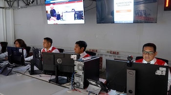 Cuatro personas con chalecos rojos sentadas frente a varias computadoras de escritorio y pantallas instaladas en la pared.