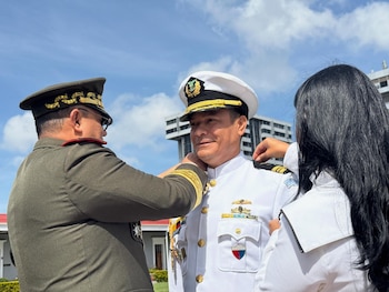 Un oficial del Ejército ajusta el cuello del uniforme blanco de un oficial de la Armada. Una tercera persona asiste al oficial de la Armada. Cielo y edificios al fondo