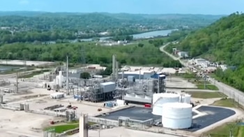 Vista aérea de un complejo industrial gris y blanco con grandes tanques de almacenamiento, tuberías y edificios, junto a un río y colinas verdes cubiertas de árboles