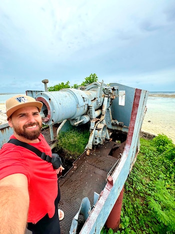 Hombre sonriente con gorra, camiseta roja y barba frente a un cañón militar gris oxidado sobre una estructura de metal en una playa, con vegetación y mar