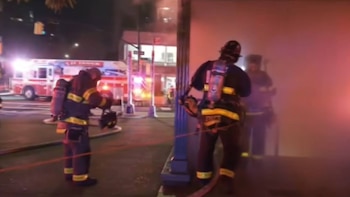 Dos bomberos con trajes oscuros y equipos de respiración en una calle. Un camión de bomberos rojo y blanco con luces encendidas visible detrás. Humo en el ambiente
