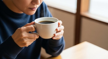 Una persona en suéter azul oscuro sostiene una taza blanca con café negro humeante con ambas manos sobre una mesa clara, fondo desenfocado.