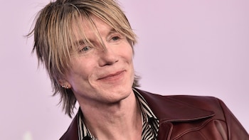 John Rzeznik, vocalista de Goo Goo Dolls, sonríe ligeramente con cabello rubio y ojos azules. Viste una chaqueta de cuero marrón rojiza sobre una camisa a rayas