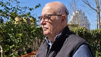 Hombre mayor con gafas, chaleco oscuro y un dispositivo de implante coclear en el lateral de la cabeza, frente a un entorno exterior
