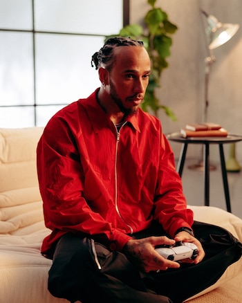 Lewis Hamilton viste una chaqueta roja y está sentado en un sofá de color claro mientras sostiene un mando de videojuegos blanco