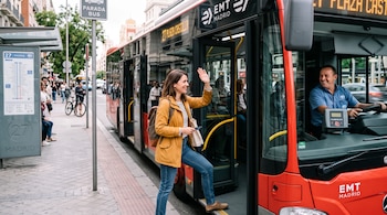 Una mujer con chaqueta amarilla sube a un autobús rojo y saluda al conductor, quien sonríe desde el volante junto a la máquina de billetes.