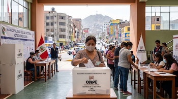Una mujer deposita un voto en una urna transparente de la ONPE en una mesa electoral con miembros de mesa y la bandera de Perú