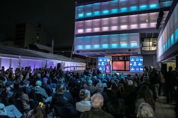 Personas sentadas de noche frente a un escenario con pantallas e iluminación azul ante el edificio del Centro Cultural Fontanarrosa