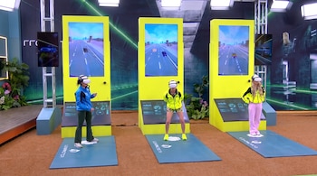 Tres mujeres con visores de realidad virtual. Tres pantallas verticales amarillas muestran un juego de autos. Se observan monitores pequeños y follaje.
