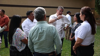 Un grupo de ocho personas, cinco hombres y tres mujeres, se reúne al aire libre. Un hombre con camisa blanca y gafas gesticula mientras habla con los demás