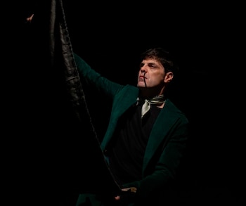 Hombre de piel clara con chaqueta verde oscuro y pañuelo claro al cuello, parcialmente iluminado contra un fondo negro, sosteniendo tela oscura