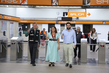 Tres personas, dos hombres y una mujer, cruzan los torniquetes de una estación de metro moderna con tarjetas en mano, bajo letreros de "SALIDA" y "TAQUILLA"