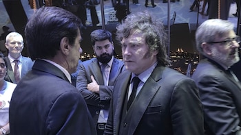 Javier Milei viste traje oscuro y habla de frente con un hombre de traje en un salón interior rodeado de personas