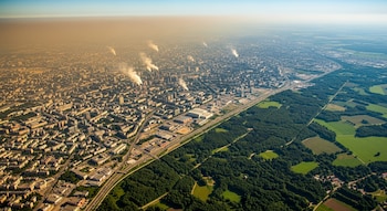 Vista aérea de una ciudad densamente construida con smog visible en el horizonte y humo saliendo de chimeneas, colindando con una zona de bosques y campos verdes.