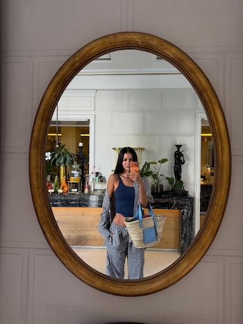 Una mujer se toma una selfie en un espejo ovalado. Viste top azul, camisa y pantalón a cuadros, y lleva un bolso de paja. Se ve un vestíbulo detrás