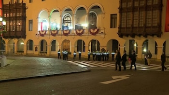 Edificio histórico iluminado con arcos, balcones de madera y adornos. Grupo de agentes de seguridad, personas caminando, calle con paso de cebra y farolas