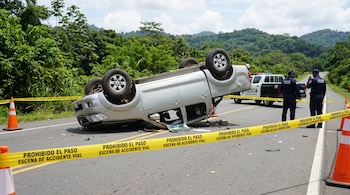 Un vehículo plateado volcado sobre el techo en una carretera, rodeado por cinta amarilla de la policía y conos de tráfico junto a dos agentes.
